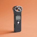 Image of a Zoom H1 black audio recorder standing upright with an orange coloured background