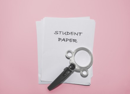 Image of a magnifying glass laying on a stack of white paper with the top piece of paper showing the text "student paper"